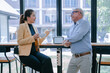 © Tj - Asian businesswoman holding a cup of coffee is engaging in a discussion with a senior businessman during a coffee break in a modern office cafeteria, fostering teamwork and collaboration