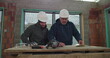 © Marco - Two construction workers closely reviewing building plans on a wooden table indoors, discussing project details, safety helmets on, teamwork and collaboration in action
