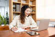 © yuttapon - Young Asian female professional wearing casual checkered shirt sitting office desk writing on paper while looking laptop screen showing engagement learning focus and positive work attitude.