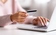 © Easy - A woman is using a laptop at home, holding a card in one hand and typing with the other. The setting is bright and cozy, indicating a casual work environment