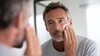 © Newleks - Mature male applying anti aging skincare product, examining facial features near bathroom mirror, emphasizing personal grooming and wellness