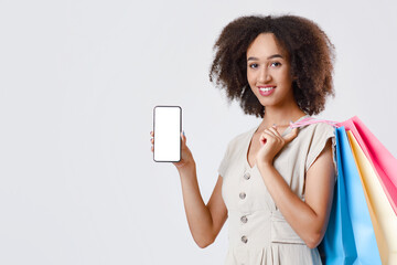 Online shopping, cashback and mobile application. Happy african american young lady holding many color packages and showing smartphone with blank screen, isolated on light background, empty space