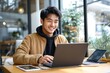 © Margarita - Young man smiling while working on laptop at a wooden table in a bright and cozy indoor space with large windows and plants outside