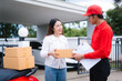 © Thitaporn - Attractive Asian delivery man in a red uniform hands a package to a middle-aged woman who is signing a clipboard next to a red motorcycle loaded with more boxes.