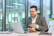 © Tetiana - Smiling young businessman in a suit sitting at his workplace in the office and talking remotely via laptop, making notes in a notebook