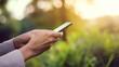 © 昌弘 嶋田 - 左手にスマートフォンを持ち、右手でタイピングする人物、早朝の光が差し込む屋外の公園、肌に柔らかく自然な影、リアルな手の質感とスマートフォンのディテールに焦点を当て、浅い被写界深度で緑の背景をぼかし、鮮やかな自然色