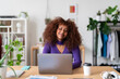 © Studio Marmellata - A young woman with curly hair is working on her laptop at a desk in a bright room. She appears focused and content, likely engaged in remote work or online learning from the comfort of her home.