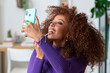© Studio Marmellata - A woman with curly hair applies lip gloss while looking at her phone. She is likely checking her makeup or recording a beauty tutorial video.