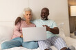 © Wavebreak Media - Diverse senior couple clicking through silver laptop on bed at home, with mug on nightstand