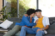 © Wavebreak Media - Laughing diverse male couple leaning together on gray couch in living room, with large glass window