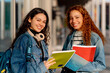 © Alberto - Two happy latin female students carrying notebooks and backpack returning to university campus