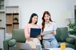 © laddawan - Happy young Asian women bonding at home on sofa, enjoying snacks, coffee, laptops, and headphones in a cozy