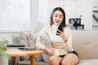© PRIME STOCK LAB - Young Asian woman sitting on sofa holding smartphone and coffee cup, smiling and relaxing in modern living room with natural light, casual home lifestyle concept