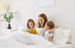 © Studio Romantic - Mother lies in bed with her two children in morning and reads book looking at illustrations. Woman is reading color book aloud, while interested kids listen attentively while lying under white blanket