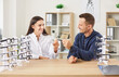 © Studio Romantic - Young smiling female optician doctor helping man patient to choose new glasses after eye examination sitting on workplace in medical clinic for vision correction. Ophthalmology and eye health concept