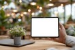 © sukarman - A person holding a tablet with a blank white screen, white shirt, natural light, blurry cafe background with out-of-focus plants 2