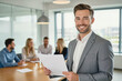© f_bossa - A confident, smiling man in a gray suit holds a document in a modern office setting with colleagues blurred in the background.