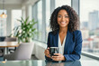 © f_bossa - A confident businesswoman with curly hair smiles warmly while holding a coffee mug in a modern office setting with a city view.