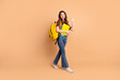 © deagreez - Young cheerful female student carrying books and a yellow backpack waving on beige background