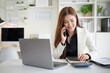 © wattana - Smiling professional woman talking on the phone while using a calculator at her desk, representing communication, productivity, and financial planning.