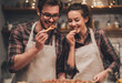© Crowded Studio - Pareja joven cocina junta en casa con delantales, compartiendo sonrisas y cariño en la cocina.Amor cotidiano, complicidad en pareja, cocina con amor, intimidad, conexión, felicidad compartida.