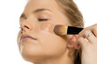 © vladimirfloyd - Close-up of young woman applying liquid foundation with makeup brush on cheek, showing smooth application process against white background