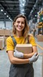 © tong2530 - Confident female worker with gloves holding package and smiling at camera in tall warehouse space, clear view of operations in background, vertical format for employer brand media