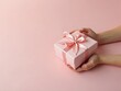 © NETI studio - Delicate pink gift box tied with a satin ribbon held by hands, on plain backdrop