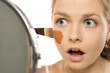 © vladimirfloyd - Young woman with shocked expression looking in mirror while applying foundation that doesn't match her skin tone, demonstrating makeup color mismatch error