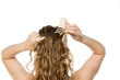 © vladimirfloyd - Rear view of young woman placing beige claw clip in her long wavy blonde hair, demonstrating half-up hairstyling process against white background