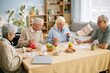 © Seventyfour - Group of senior friends eating breakfast in retirement home sitting around table, sharing stories and laughter. Table set with fruit and pastries, surrounded by cozy, homely atmosphere