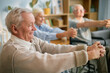 © Seventyfour - Elderly people in retirement home performing stretching exercises while seated, focusing on health and well-being amid light-filled indoor setting with plants and bookshelves