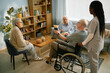 © Seventyfour - Group of elderly individuals sitting together in bright room, engaging in conversation and activities, with caregiver assisting. Bookshelf and plants in background