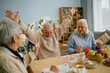 © Seventyfour - Group of elderly friends enjoying each other's company while playing a board game around dining table, with cheerful expressions and raised hands in celebration