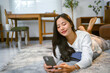 © Crystal - Asian woman lying on a cozy carpet in a modern living room, enjoying leisure time while browsing on her smartphone and relaxing at home
