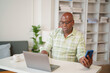 © MINAE - Senior African American man wearing casual clothes and glasses, working from home at a desk with a laptop and smartphone, focused in a living room setting
