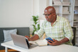 © MINAE - Senior african american man is sitting at his desk, working from home, using a laptop and smartphone, taking notes in a notebook and holding a pen