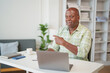 © MINAE - Cheerful african american businessman pointing finger to laptop screen during online meeting while sitting at desk in home office, he is explaining something to his colleagues