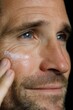 © serdon - A close-up shot captures a bearded young man as he applies face cream and looks directly at the camera