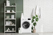 © Pixel-Shot - Modern washing machine with shelving unit, houseplant and detergents near white brick wall in laundry room