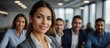 © ULTRA STOCK'S - Confident and poised Latin businesswoman stands in office with team working behind her.