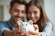 © Yuliia - A young couple smiles at each other while making a joint commitment to saving and budgeting by inserting coins into a piggybank in close-up shot.