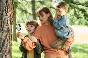 Wall Mural - Waist up portrait of mother with two boys holding magnifying glass while exploring nature together in green forest