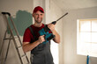 © Grustock - Cheerful construction worker in overalls and a red cap is holding a power drill in a partially painted room, standing beside a ladder near a sunlit window