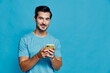 © SHOTPRIME STUDIO - Smiling young man wearing a blue t-shirt holds a smartphone, looking at the camera with a friendly expression, isolated against a solid light blue background.
