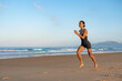 © sutulastock - Focused athletic woman doing a forward lunge jump on a sandy beach at sunrise, mid-air with graceful form, surrounded by ocean waves and distant mountains in golden morning light.