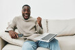 © SHOTPRIME STUDIO - Cheerful young man celebrating success while working on a laptop at home, wearing a cozy sweater in a minimalist living room setting