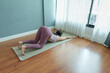 © eggeeggjiew - Asian woman performs a extended puppy yoga pose stretching her arms and back in a room with large windows and wooden flooring.