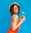 © Prostock-studio - Happy black girl in swimsuit enjoying exotic summer coconut cocktail and smiling at camera, posing over blue background in studio, copy space