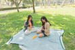 © WMSTUDIO - Two Asian Women Enjoying a Relaxing Afternoon in a Park, Playing Ukulele, and Sharing Joyful Moments on a Green Blanket Surrounded by Nature